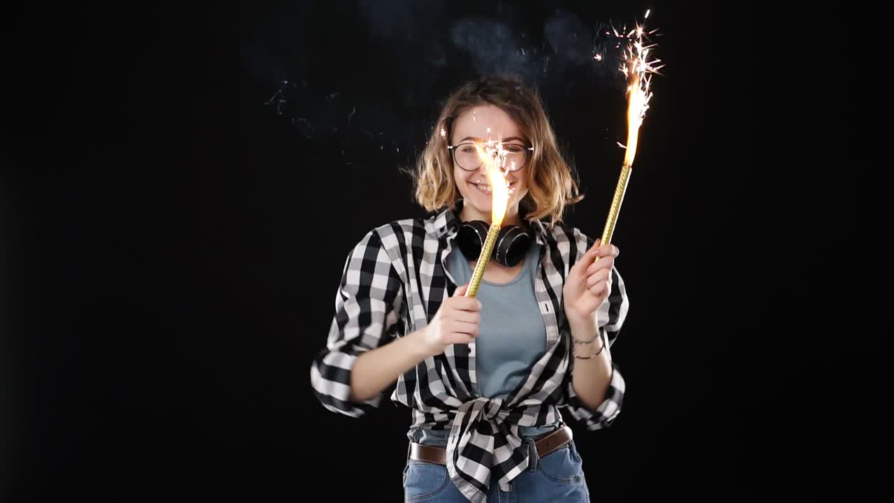 European hipster girl in black and white plaid shirt, headphones on neck celebrating with sparklers and bengal fire. Happy young woman having fun and playfully dancing against black background. Slow motion