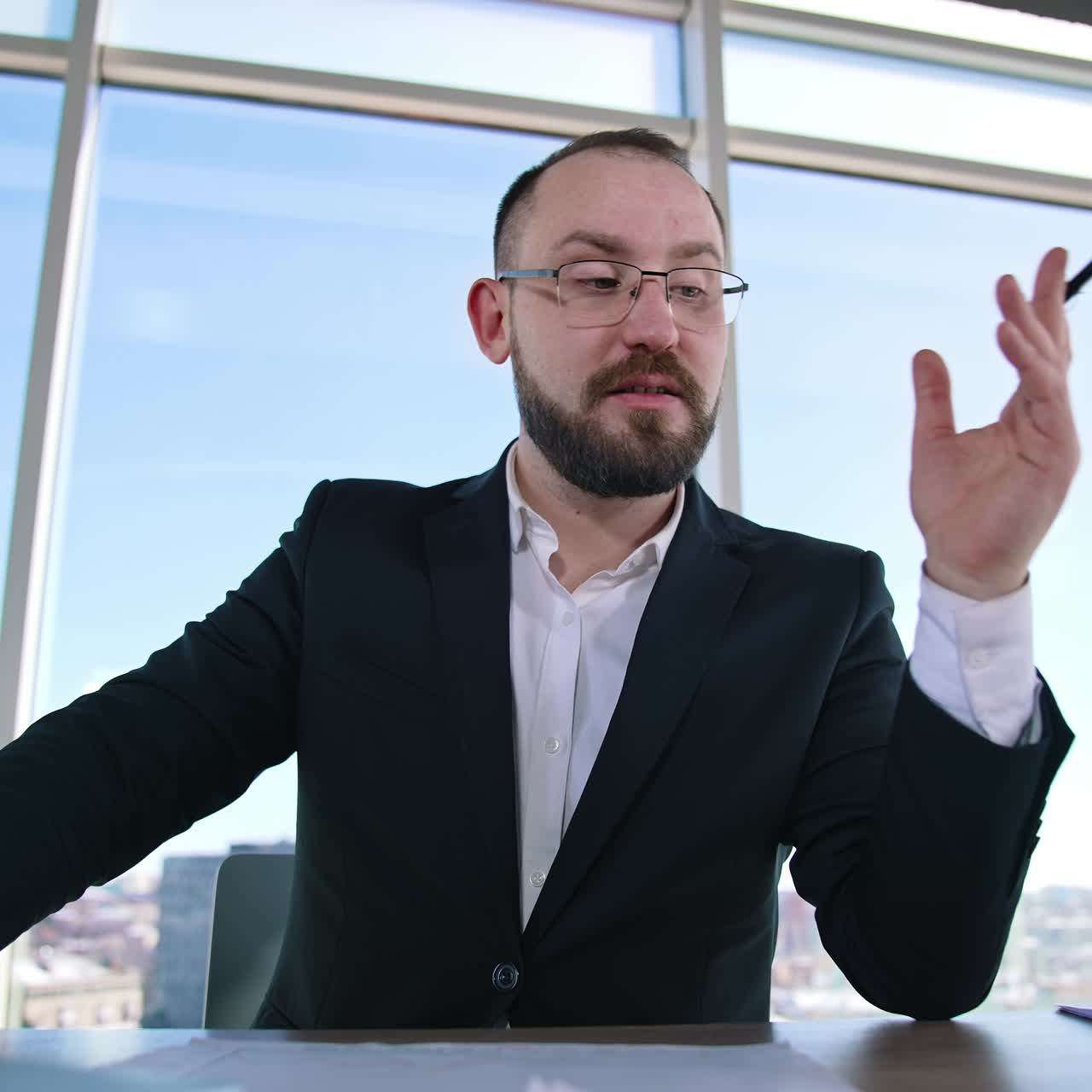 Portrait of a businessman sitting in office. Bearded entrepreneur in suit and glasses talking about business on window backdrop