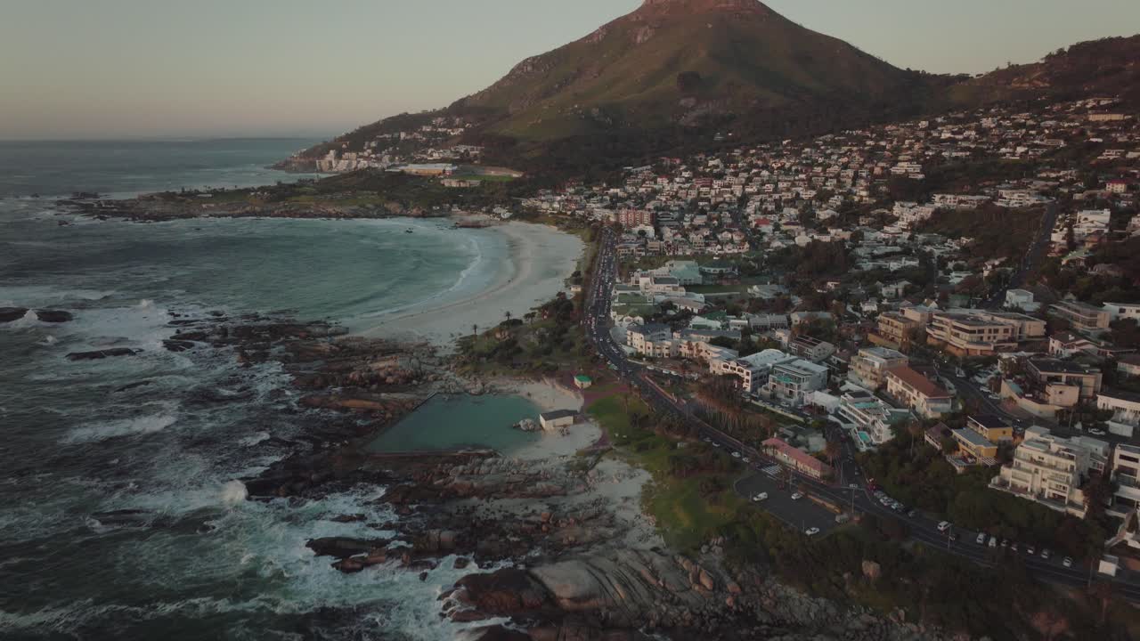 el dron se inclina hacia arriba y vuela alto sobre la playa de camps bay en ciudad del cabo, sudáfrica. en el fondo, la montaña de la cabeza de león se eleva iluminada por la puesta de sol.