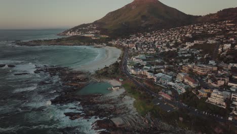 El-Dron-Se-Inclina-Hacia-Arriba-Y-Vuela-Alto-Sobre-La-Playa-De-Camps-Bay-En-Ciudad-Del-Cabo,-Sudáfrica;-Al-Fondo,-La-Montaña-De-La-Cabeza-Del-León-Se-Eleva-Iluminada-Por-La-Puesta-De-Sol.