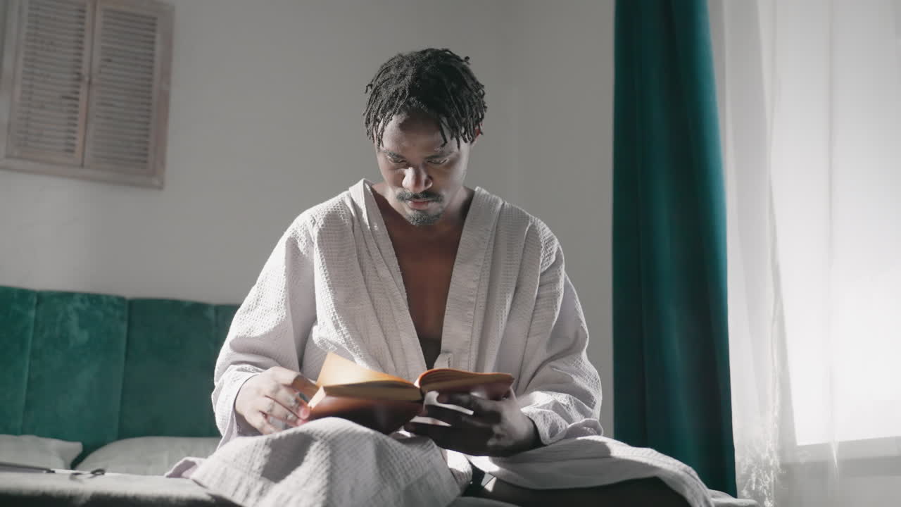 Guy in bathrobe sitting on bed, reading a book in morning light. Quiet, cozy room with relaxed atmosphere. Soft sunlight highlights focused expression while reading, enjoying peaceful solitude