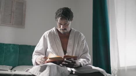 guy in bathrobe sitting on bed, reading a book in morning light. quiet, cozy room with relaxed atmosphere. soft sunlight highlights focused expression while reading, enjoying peaceful solitude