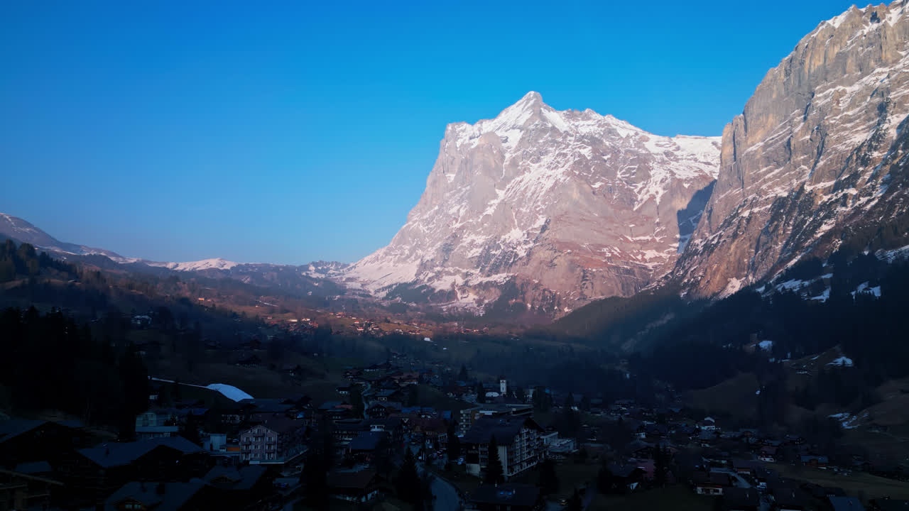 el dron asciende mostrando el fresco pueblo de grindelwald, suiza, en la base de las nevadas montañas grises.
