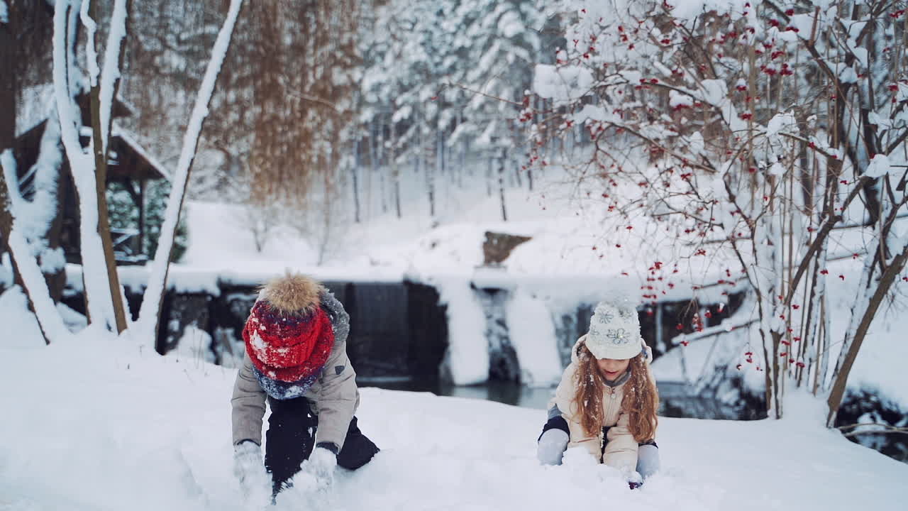 Happy children wearing warm coats and caps are throwing snow up on the snowy background. Little kids are playing with white snow near the beautiful river in the forest. Slow motion.