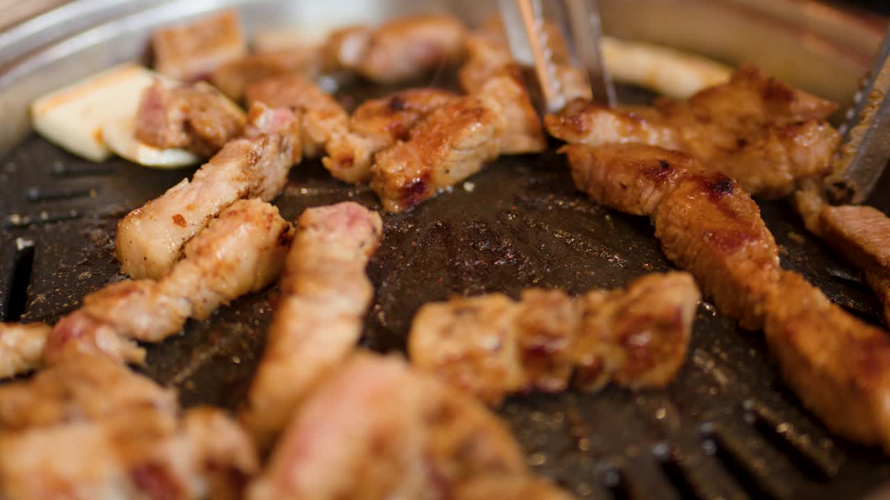 Close-up of pork belly grilling on tabletop pan as metal tongs turn sizzling pieces
