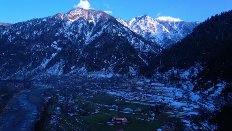 Aerial-view-of-a-river-and-village-on-the-side-with-snowy-peaks-in-the-background-in-the-evening