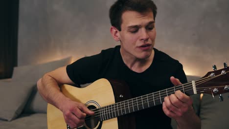 Portrait-of-a-cheerful-man-singing-a-lyric,-emotional-song.-Playing-on-an-acoustic-guitar-and-enjoying-it,-while-sitting-on-a-grey-couch-with-a-backlight-under.-Slow-motion