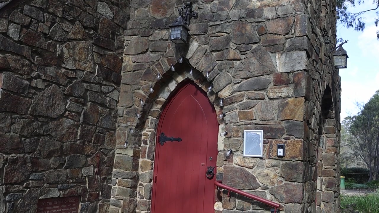 Olinda, Melbourne'da çarpıcı kemerli kırmızı kapısı, dokulu rustik taş işçiliği, doğal dış mekan ışığı ve sakin yeşil çevresiyle eski bir taş kilisenin geniş çekimi. İnsan görünmüyor.