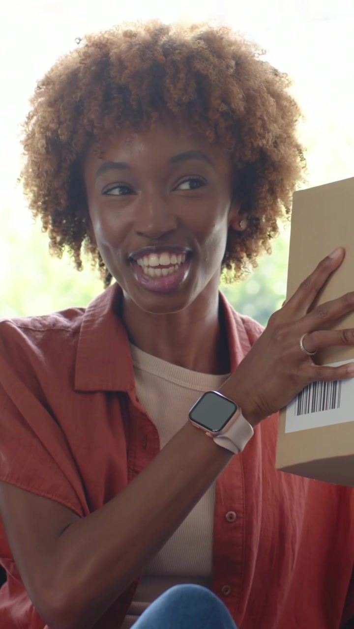 Tenendo una scatola di cartone verso la telecamera, donna con maglietta color ruggine a casa che ispeziona il codice a barre preparando l'unboxing