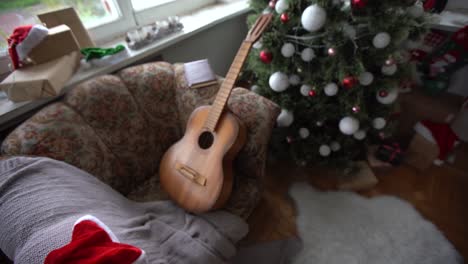 un anciano de primer plano con el tradicional traje rojo de papá noel con barba blanca se sienta cerca de un árbol de navidad decorado, toca la guitarra, concepto de felicitaciones, feliz navidad.