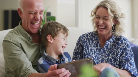 Glückliche-Kaukasische-Großeltern-Mit-Enkel,-Der-Tablet-Benutzt-Und-Im-Wohnzimmer-Sitzt