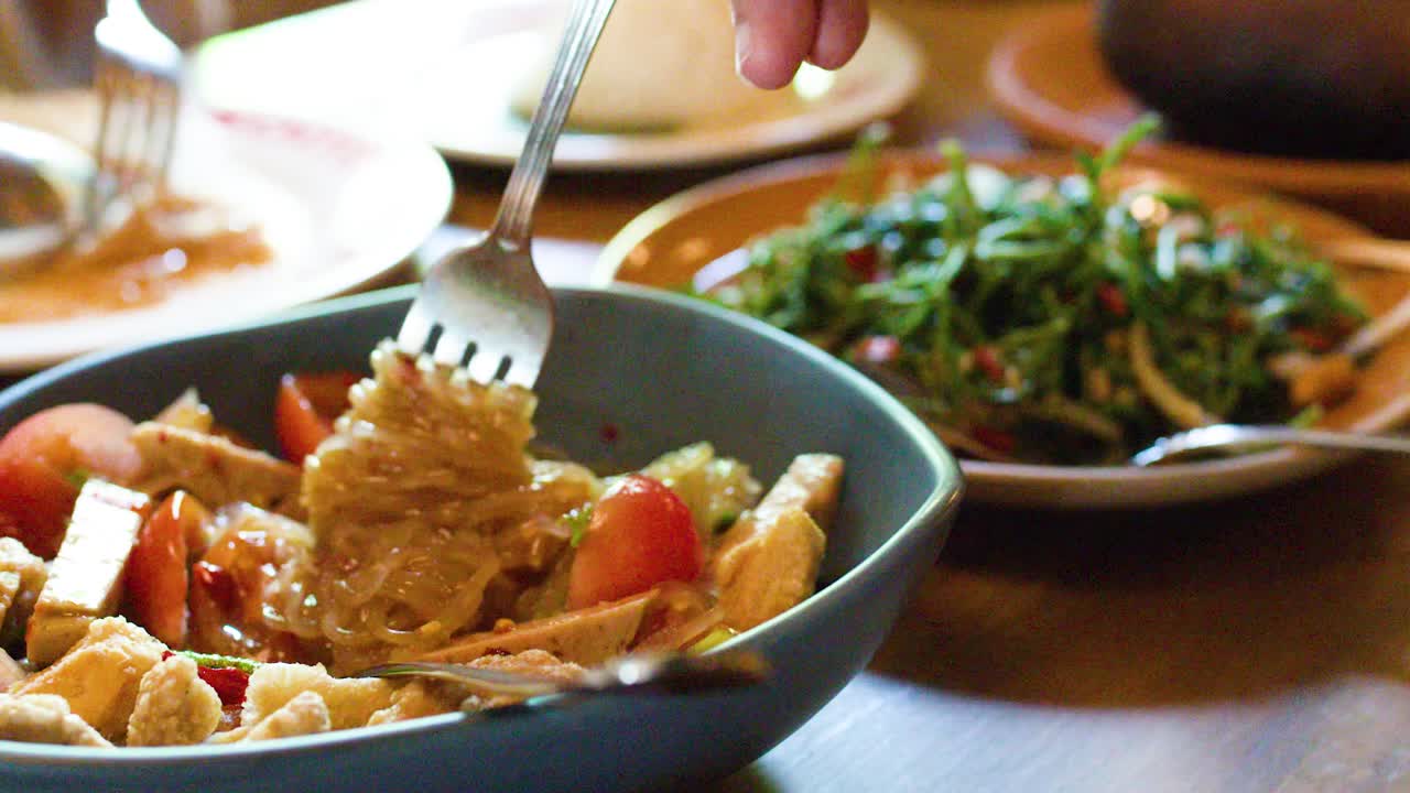 Hand twirls rice vermicelli salad with tofu, tomato, and chili in warmly lit Thai restaurant