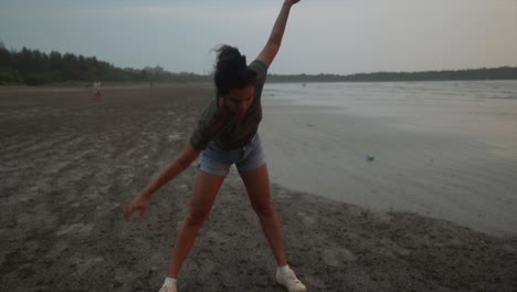 Landscape-view-of-a-young-woman-doing-stretching-exercises,-on-a-sandy-beach-by-the-ocean,-on-a-cloudy-day