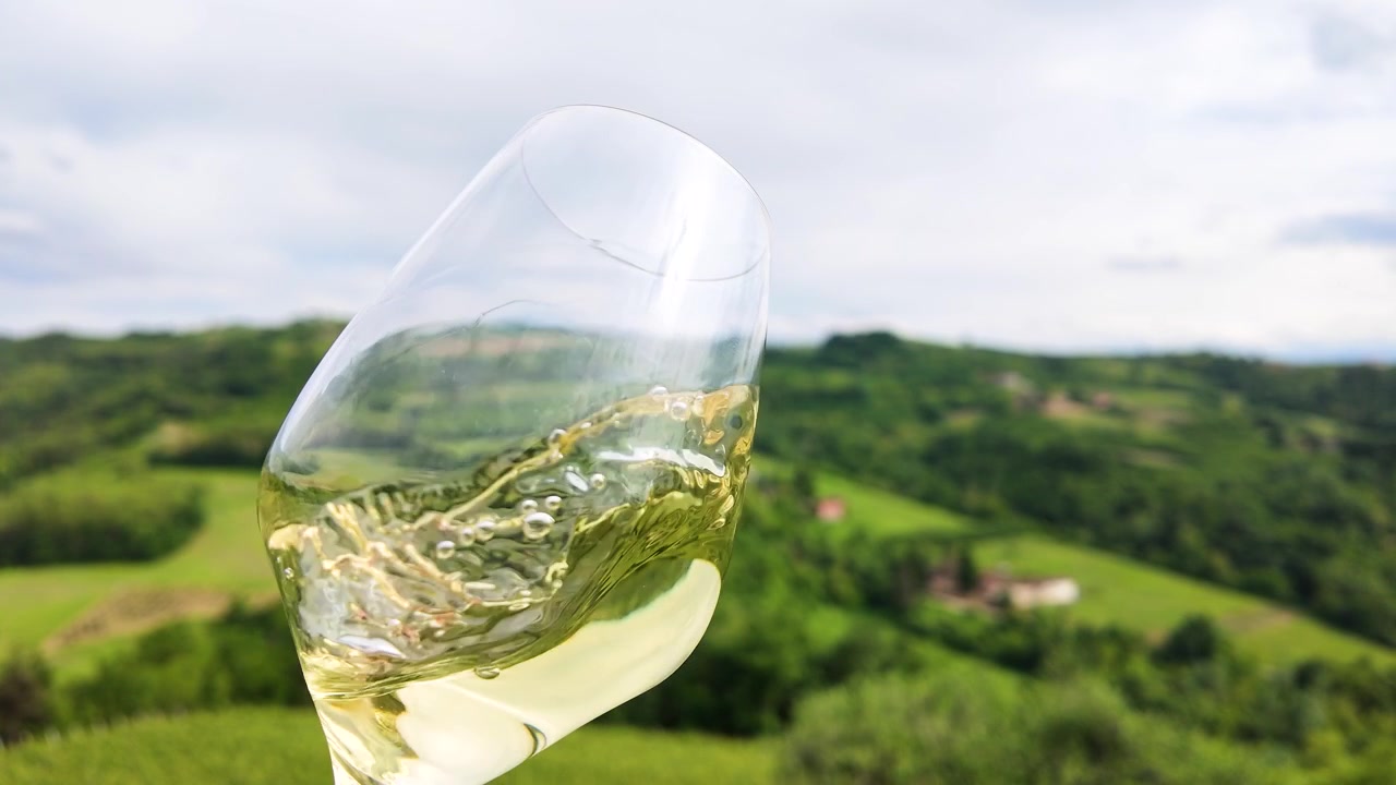 Primo piano di vino bianco che volteggia in un bicchiere, sullo sfondo di un vigneto panoramico con dolci colline.