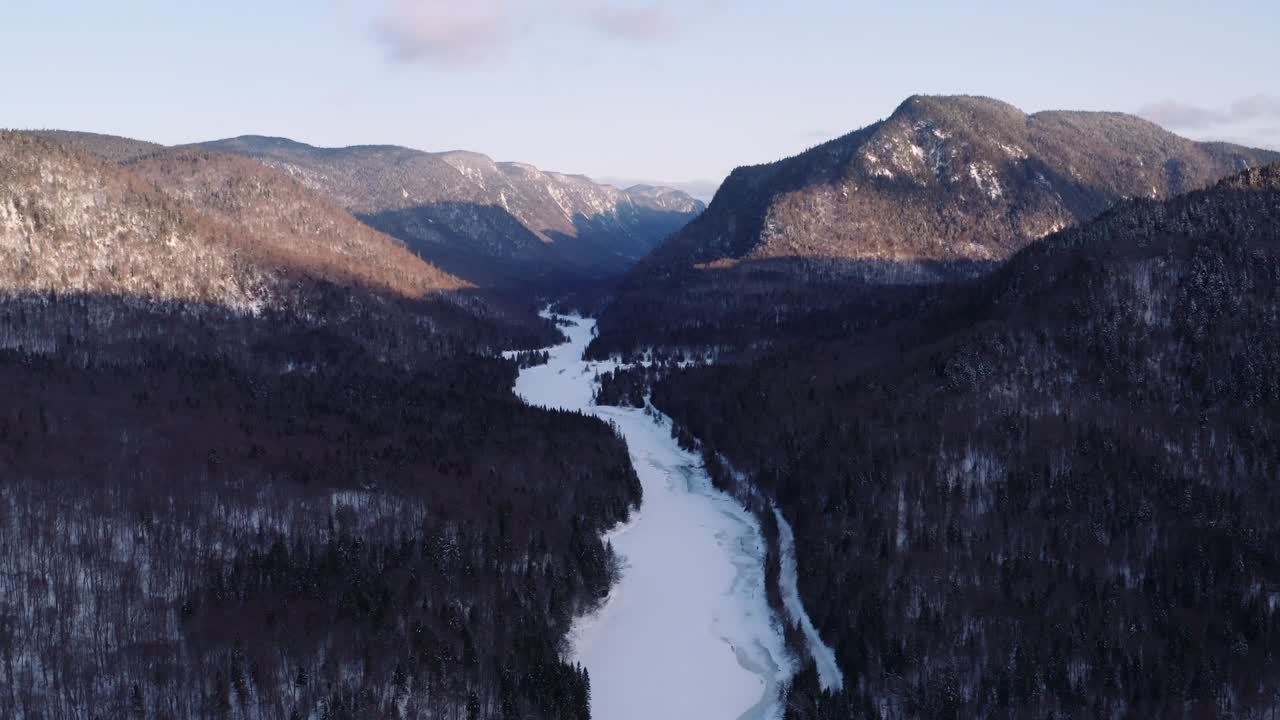Episk bagudbevægende vidvinkel luftoptagelse af landskab med en sneklædt flod, der løber gennem en fjord og ind i en vinterborealskovdal.