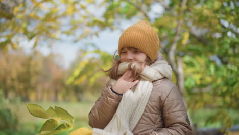 side view of cute girl smiling warmly wearing mustard beanie beige quilted jacket and thick scarf while holding scarf gently in hand surrounded by vibrant green and yellow foliage