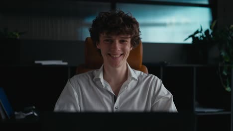 Portrait-of-a-happy-young-guy-with-curly-hair-working-at-a-laptop-posing-and-smiling-in-the-office