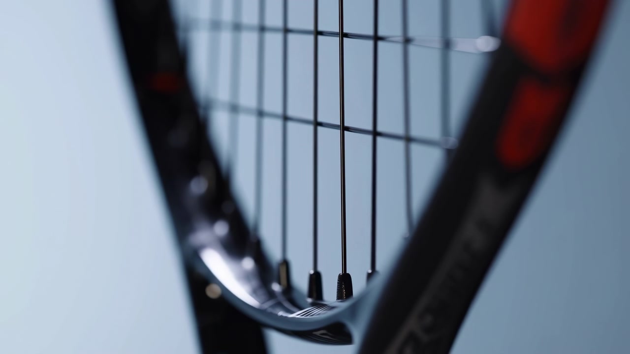 Detailed view follows lower section of strung racket as frame drifts gently across composition, with crisp vertical strings, glossy black surfaces, soft highlights, and blurred blue backdrop.