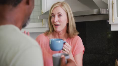 vista sobre el hombro de una pareja mayor diversa bebiendo café y hablando en la cocina
