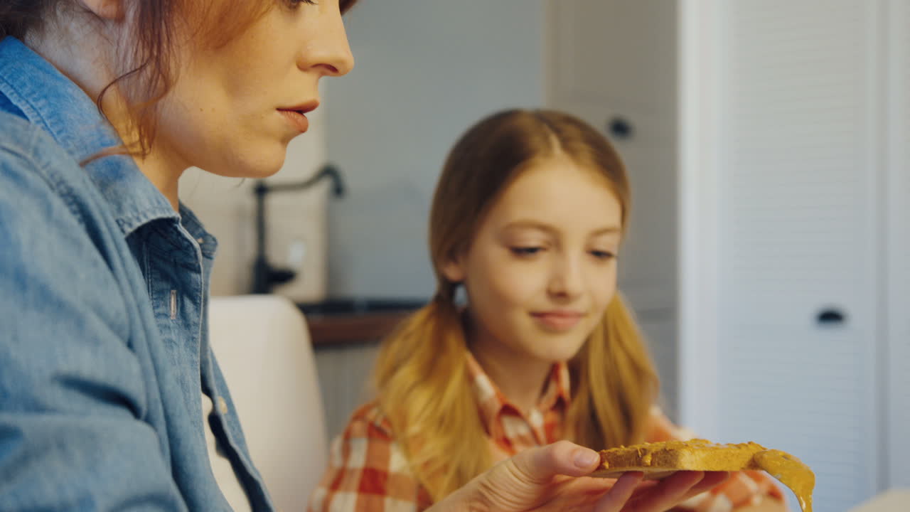 cerca de la madre bonita extendiendo mantequilla de maní en el pan con una cuchara para su hija mientras la observa en la cocina. adentro