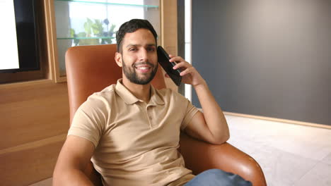 smiling man sitting in chair, talking on smartphone in modern office
