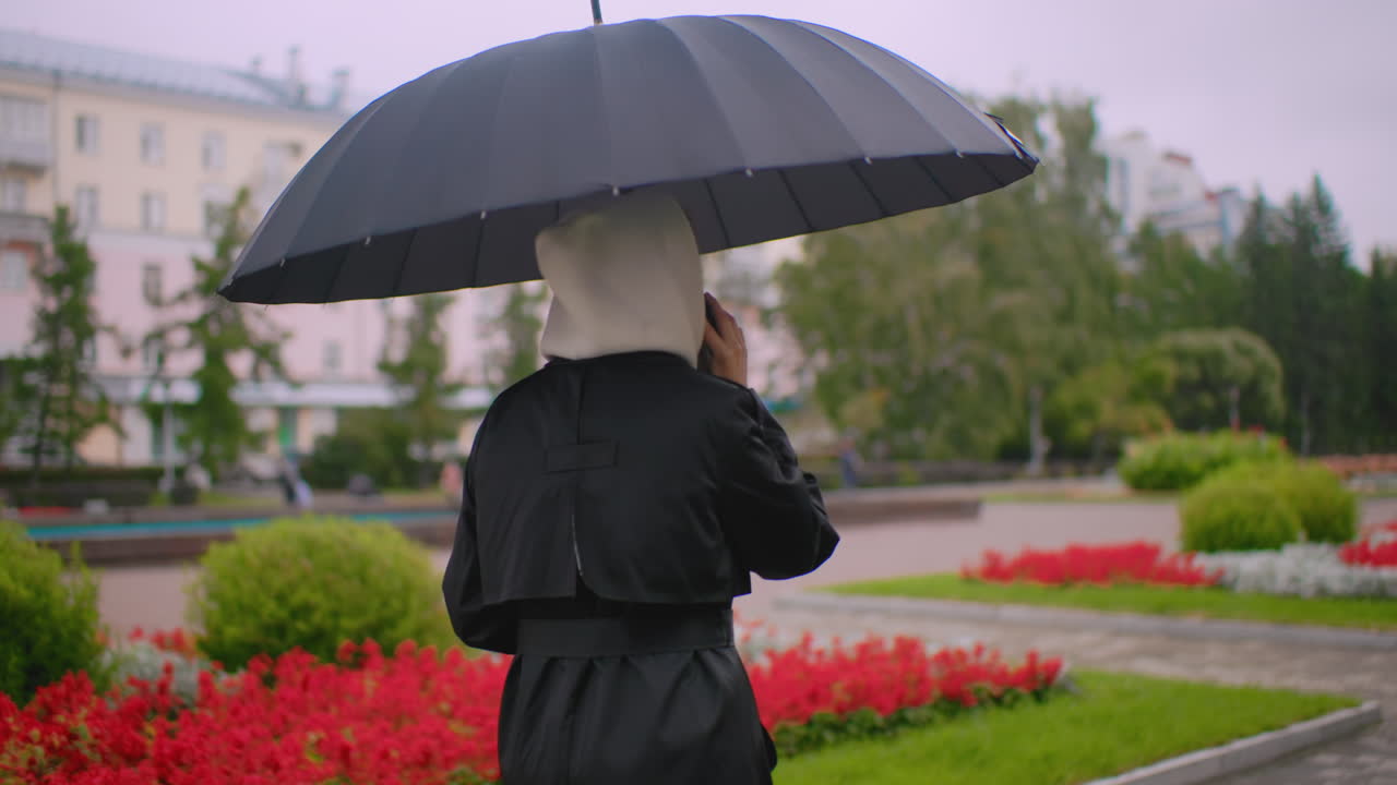 Back shot of a female in black coat with hood walks under umbrella on rainy park path, answering call, surrounded by red and white flowers, green trees and cloudy sky creating calm urban rainy day mood