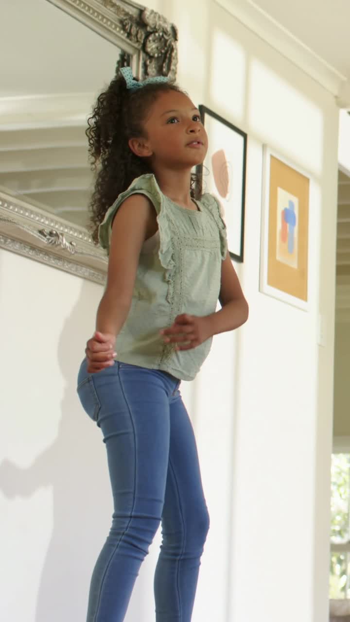 Vertical video: Off-camera cue causing girl with blue bow green top dancing at home near mirror art