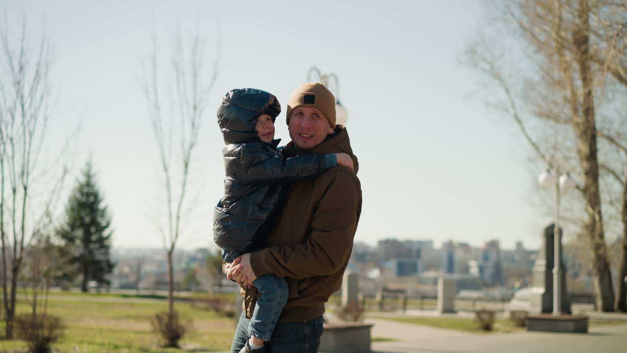 Father carrying his son, the child is dressed in a shiny black puffy jacket, and the man is wearing a beanie,with a blurred cityscape and open field in the background