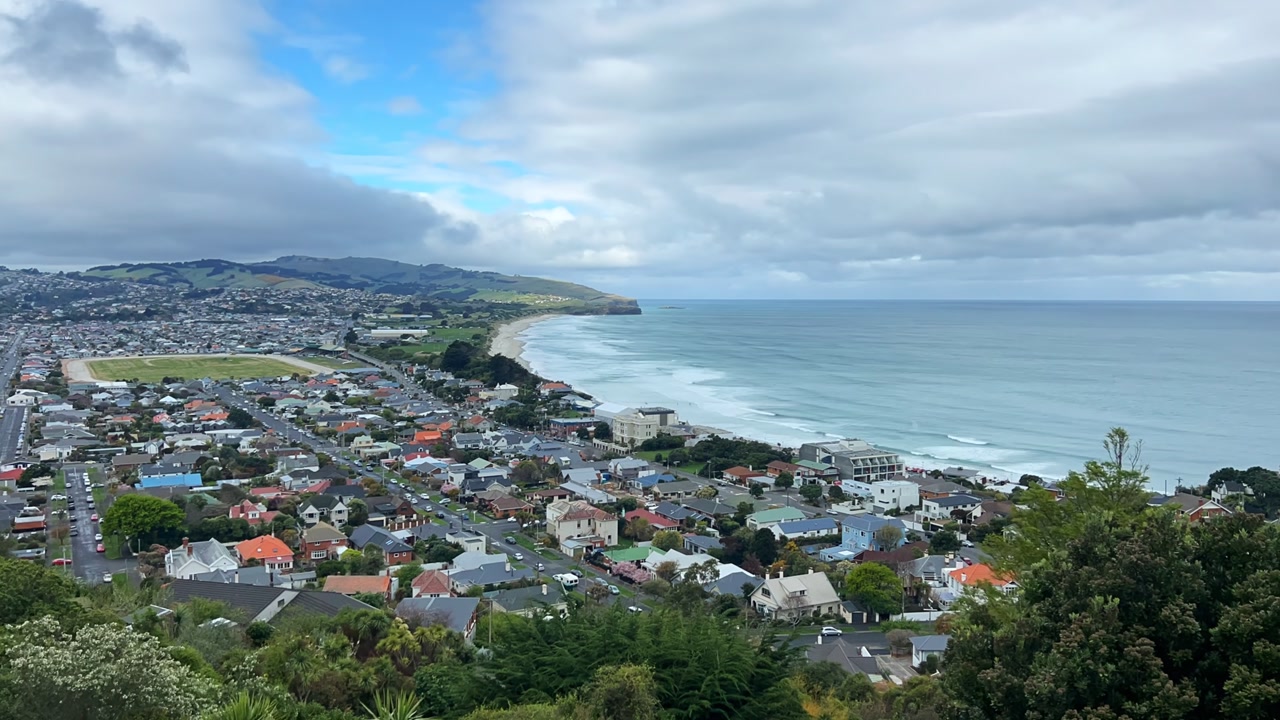 Wide elevated time lapse over Dunedin's southern suburbs and ocean beach. Residential streets meet rolling surf under partly cloudy skies.