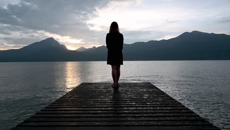 Girl-standing-at-the-end-of-the-pier-admiring-the-beautiful-sunset,-lake-and-the-mountains