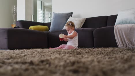 bebé caucásico jugando con una taza de plástico mientras está sentado en el suelo en casa