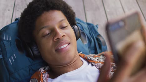 Smiling-african-american-man-in-city,-lying-on-deck-wearing-headphones-and-using-smartphone-in-park