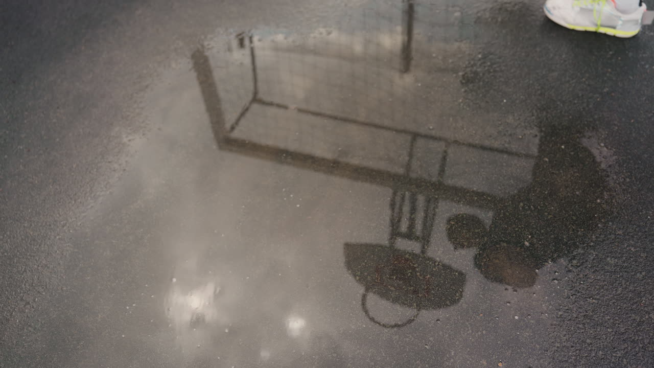 Pfützenreflexion eines Fußballtors, zwei Silhouetten unter einem Regenschirm auf feuchtem Asphalt, filmischer, stimmungsvoller Regen mit glitzernder Oberfläche und subtilen Wellen, entfernte Stadionlichter und nostalgische Atmosphäre