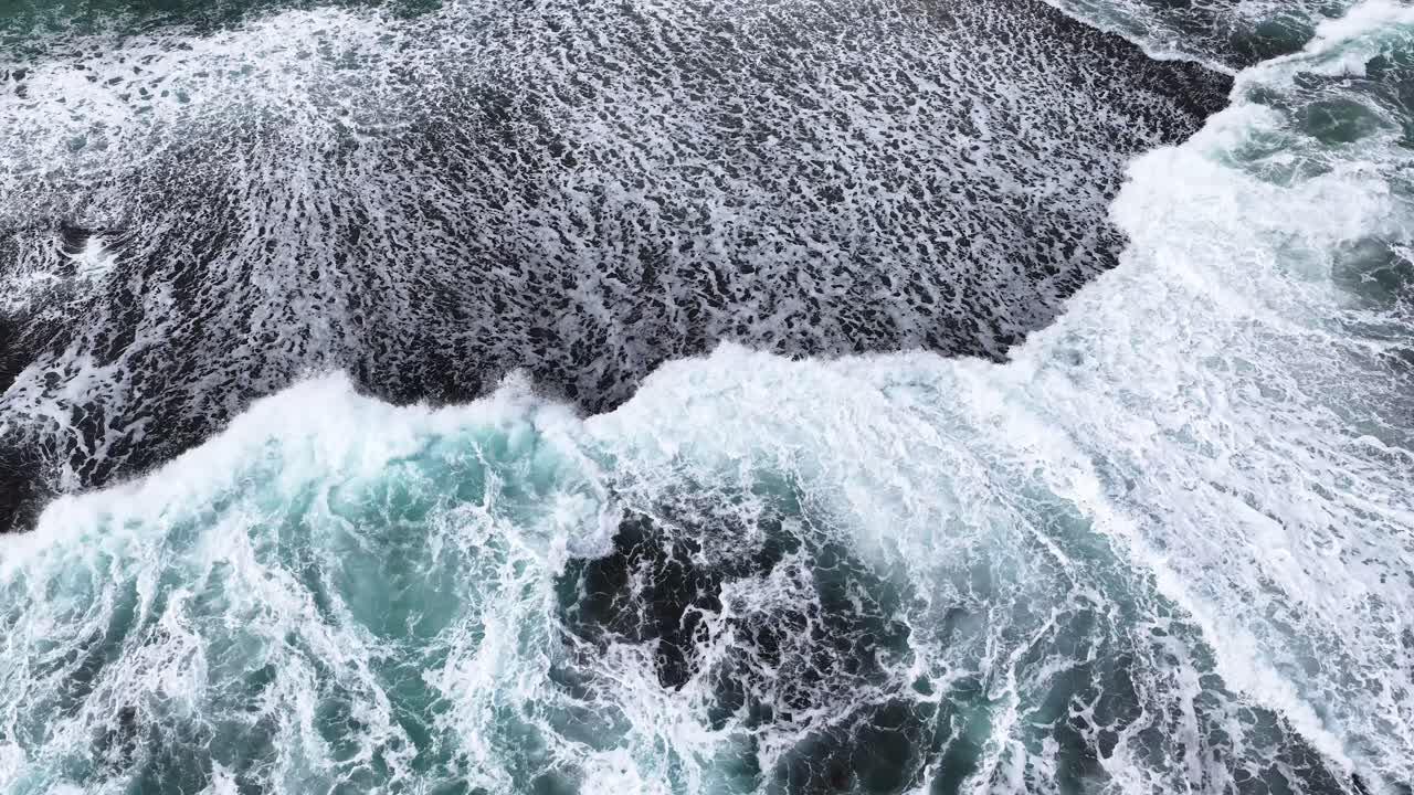 Overhead drone shot of foamy waves breaking on dark sand, natural daylight, smooth camera movement
