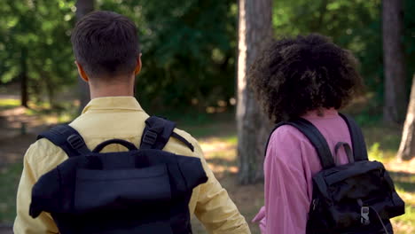 hiker couple with backpacks walk on a forest trail.young black female and caucasian man holding hands. view from behind.