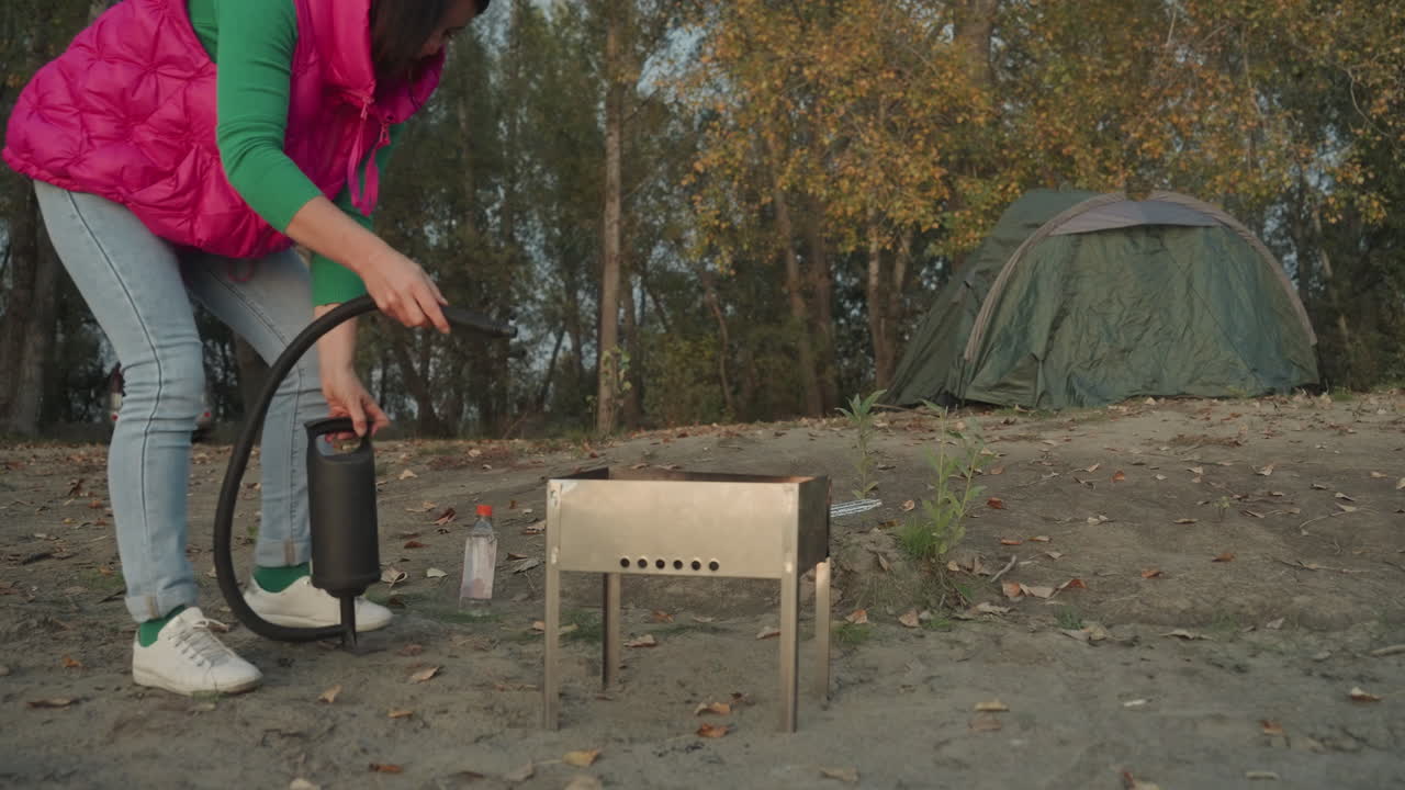 Kvindelig campingopsætning. En person forbereder et bærbart komfur ved skovbrynet i efteråret. Kvinden samler omhyggeligt campingkomfuret nær sit telt, omgivet af den rolige skovnatur i efteråret.