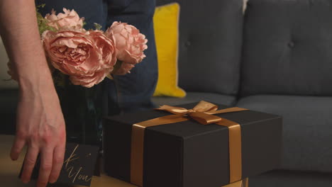 close up of man putting gift wrapped present with thank you card and flowers on table in lounge at home