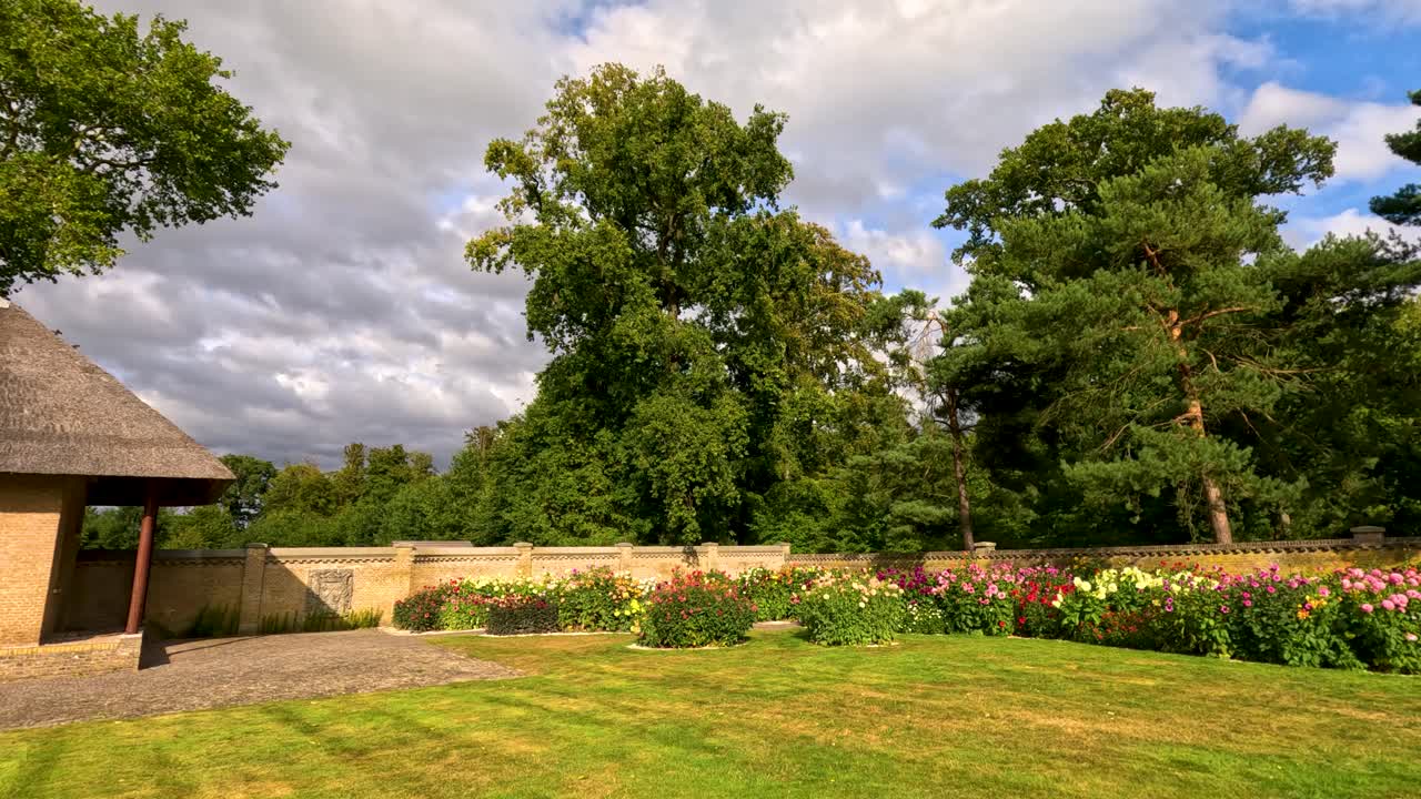 A smooth daytime pan reveals a charming stone cottage, vibrant dahlia flower beds, and lush greenery under partly cloudy skies in a botanical garden