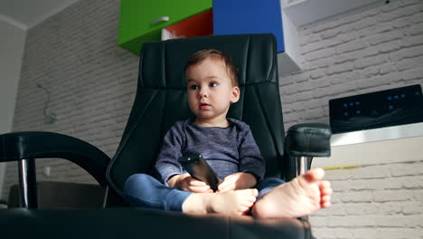 calm caucasian child sitting in a comfortable office holding remote control. lovely boy looks away and smiles adorably to approaching camera. low angle view close up.