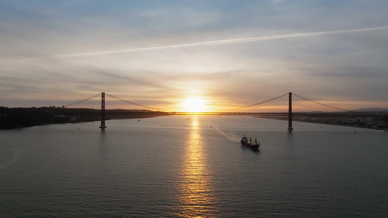 Rivelazione lenta con drone del Ponte 25 de Abril in silhouette, Lisbona al tramonto. Il sole si riflette sul fiume Tago. Navi che passano sotto. Filmato aereo cinematografico del Portogallo.