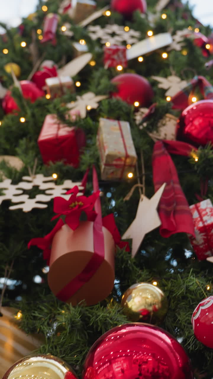 Close-up of Christmas tree with festive ornaments, golden baubles, red ribbons, decorative squirrel, and twinkling lights shining through green branches in modern shopping mall setting