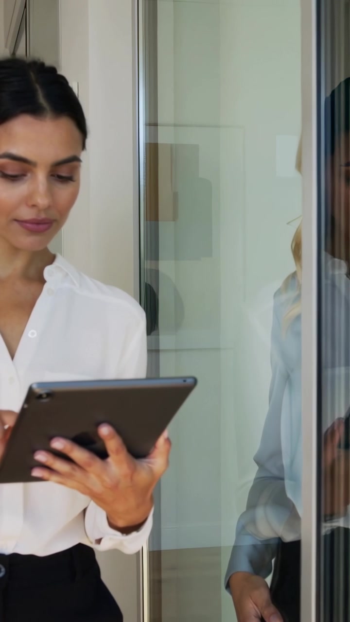 Video verticale: Donna in bianco che usa un tablet vicino a una parete divisoria in vetro dell'ufficio, con un collega che revisiona.