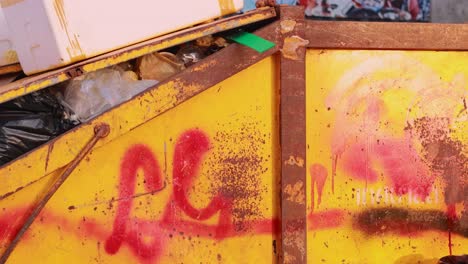 a yellow dumpster with graffiti overflows with trash in a phuket alley, captured in natural daylight