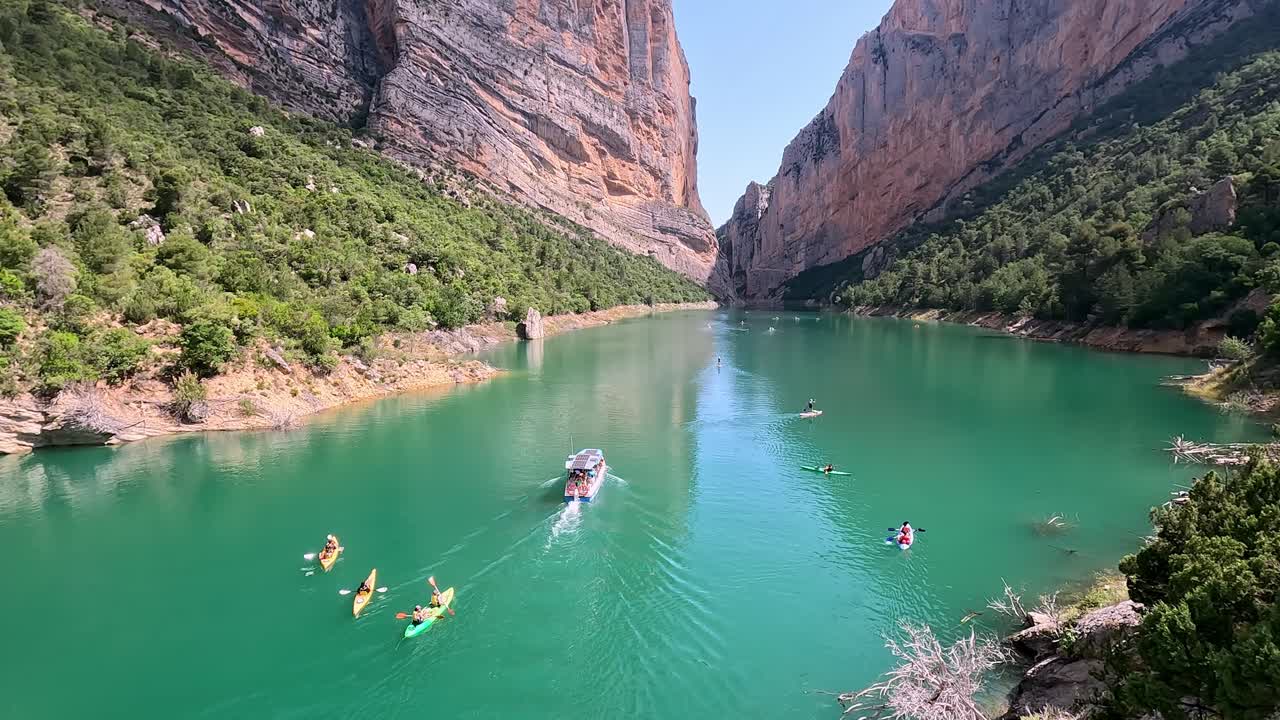 Bright, sunny day with kayakers paddling and a tour boat navigating the turquoise waters of Congost de Mont-rebei. Surrounded by dramatic cliffs and lush greenery.