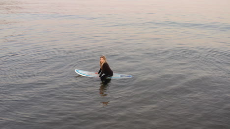 drone shot of woman wearing wetsuit sitting on surfboard at sea