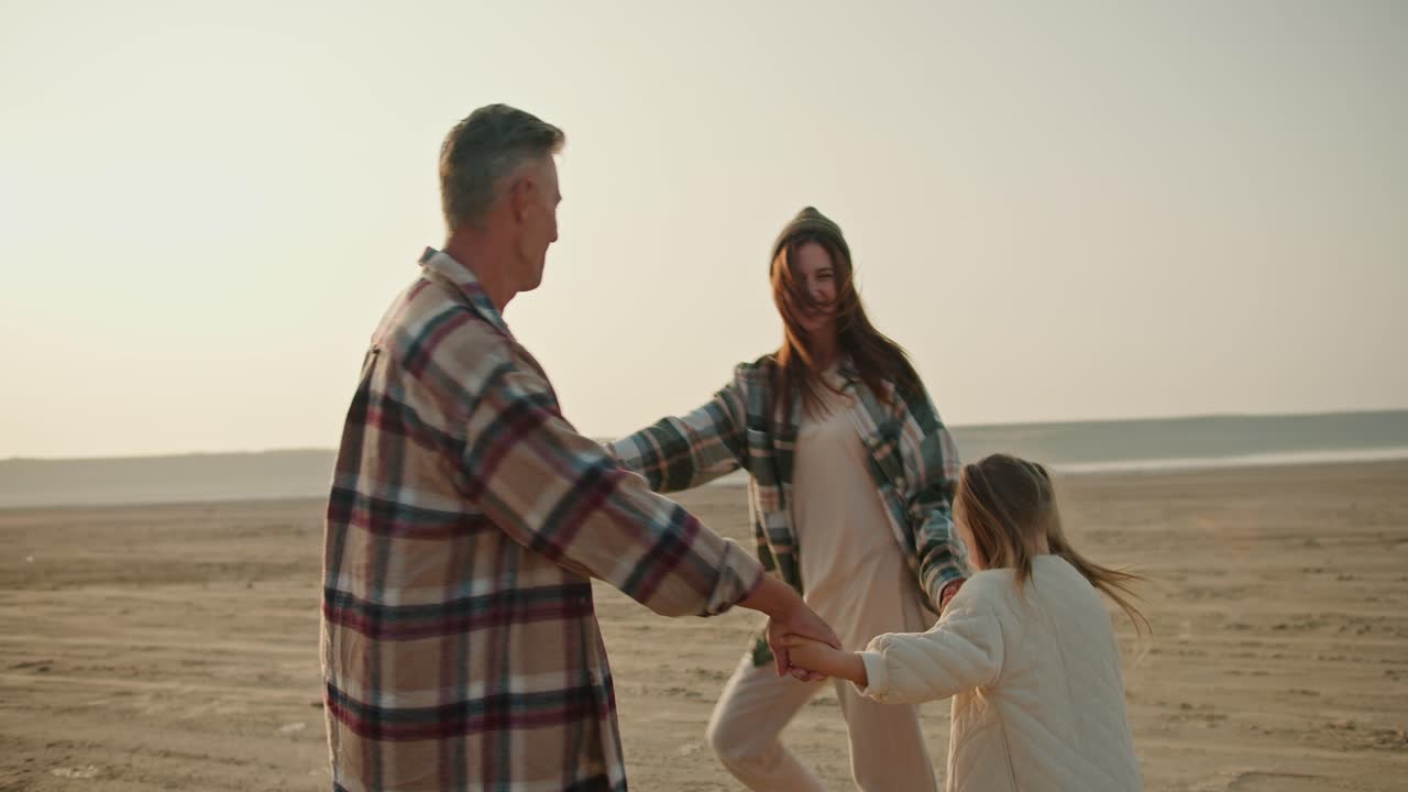 가족의 행복한 트리오, 체크 셔츠를 입은 갈색 남성, 초록색 체크를 입은 아내와 색 재을 입은 작은 딸과 함께, 사막 해변에서 춤을 추고, 도시 밖의 피크닉과 휴가 중에 원을 고, 원을 춤을 니다.