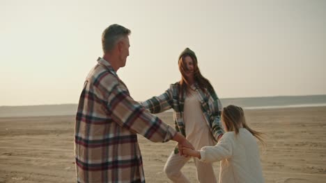 A-happy-trio-of-a-family,-a-brunette-man-in-a-checkered-shirt,-together-with-his-brunette-wife-in-a-green-checkered-shirt-and-a-little-girl-daughter-in-a-white-jacket,-dance-on-a-deserted-seashore-and-spin-in-a-circle-and-dance-in-a-circle-during-their-picnic-and-vacation-outside-the-city