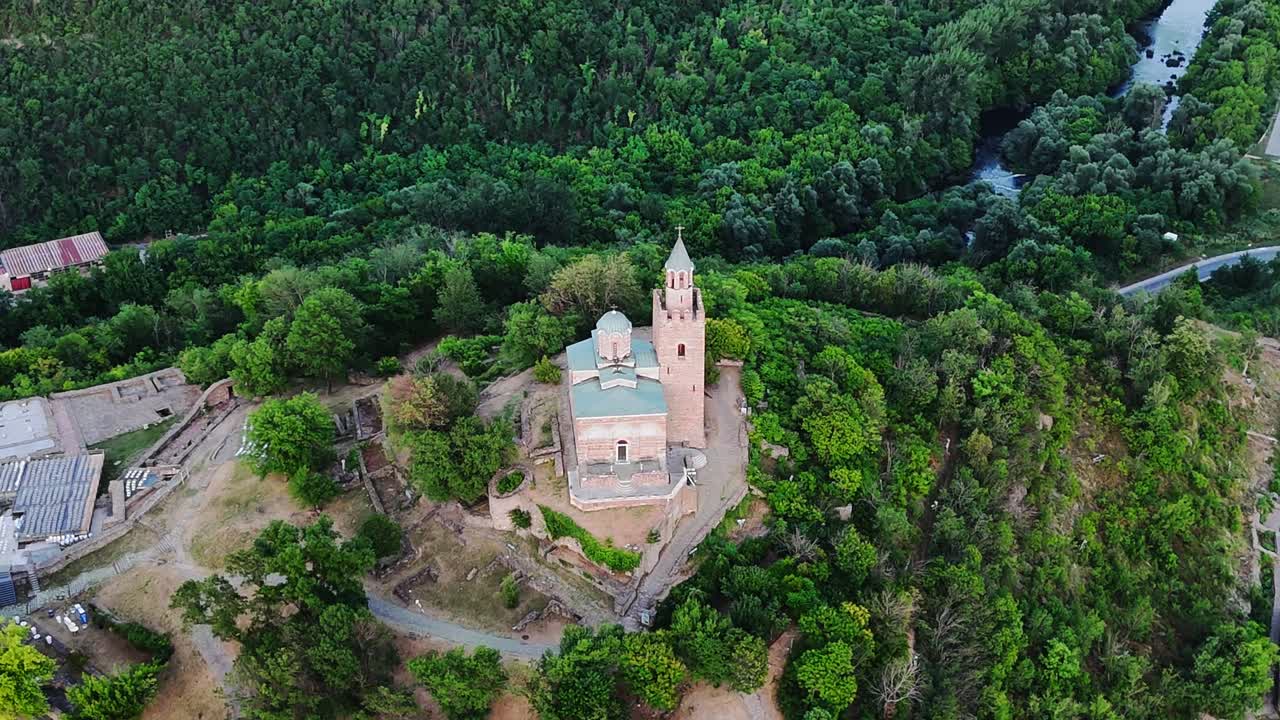 Ripresa con drone in orbita laterale che si avvicina alla chiesa in cima alla Fortezza di Tsarevets, con alberi verdi, tetti della città vecchia e il fiume Yantra in basso