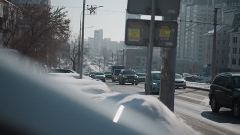 High angle winter city scene with cars moving through two lanes while people walk along pedestrian path, snowbanks lining road, distant towers, cold daylight haze, and commuting rhythm