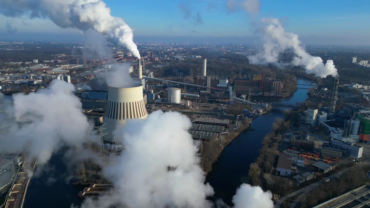 Aerial view of a thermal power plant emitting smoke on a sunny day, with a river and a city in the background. Majestic aerial view flight drone shot footage from above